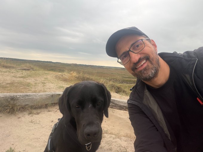 A smiling man with glasses and a cap takes a selfie with a large black dog on a sandy beach with grassy dunes and an overcast sky.