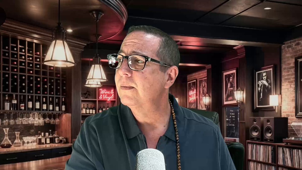 The image features a man wearing glasses, sitting at a bar with a microphone in front of him. He appears to be talking into the microphone or recording something. The background includes several wine bottles displayed behind the man, suggesting that he might be discussing topics related to wine or hosting a podcast about wine.