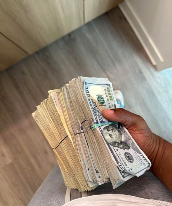A hand holds a thick stack of banded $100 bills, the clearly visible currency featuring Benjamin Franklin's portrait and the "100" denomination. The image visually reinforces the post's message about relentlessly "grinding for the money," showcasing a large sum of cash as a symbol of financial hustle.