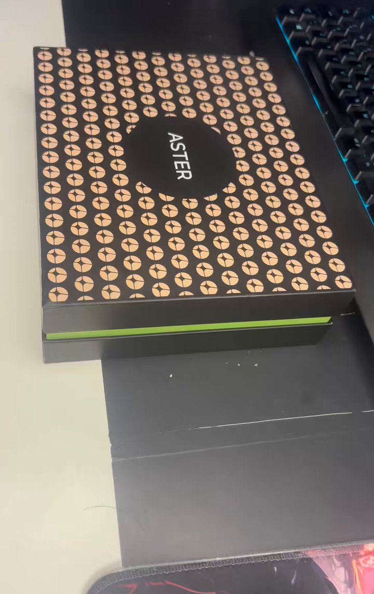 The image shows a black box with a brown design, sitting on top of a desk next to a keyboard. The box has the word "ASTER" written on it in white letters. There are also some text elements visible in the image that read "$ASTER unbox".