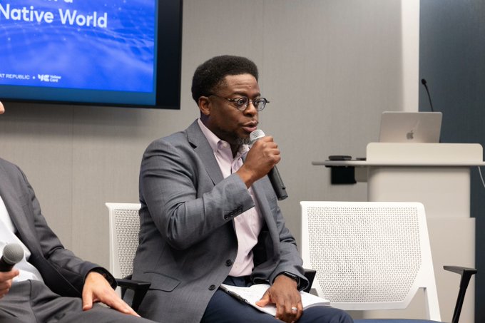 A man in a gray suit and glasses speaks into a microphone while seated on a panel, with a screen behind him displaying 'Native World' and 'NE... GREAT REPUBLIC'.