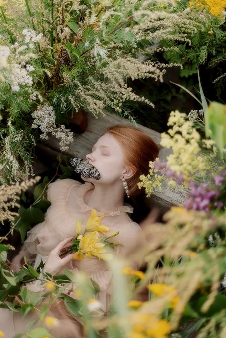 An artistic photograph of a woman with red hair and a butterfly over her mouth, surrounded by lush green foliage and yellow flowers.