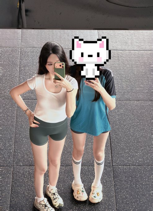 Two women pose for a mirror selfie at what appears to be a gym, one wearing a white fitted top with dark green shorts and the other in a blue athletic top with white knee-high socks and crocs, with a pixelated white bunny sticker covering her face. The caption playfully frames the workout session as being "supervised" by the friend (likely the woman on the left) acting as a "teacher" at the gym.