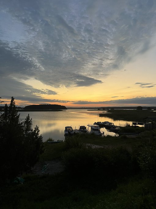 A serene lake scene at sunset, with boats docked along the shore beneath a dramatic sky filled with textured clouds and warm golden light reflecting on the calm water. The peaceful "gmeme" (good morning meme) captures a tranquil start to the day with its breathtaking natural beauty.