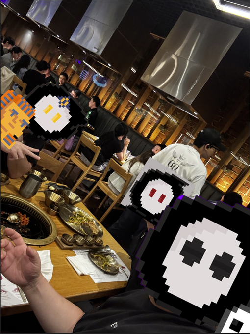 A view from inside a busy restaurant where people are seated around a table with hot pot dishes and side plates, with pixel-art ghost and skull stickers overlaid on the image.