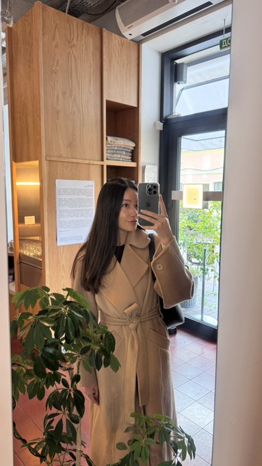 A mirror selfie of a young woman with long dark hair wearing a tan trench coat, holding a phone in what appears to be a room with wooden furniture, plants, and a glass door leading to a balcony.