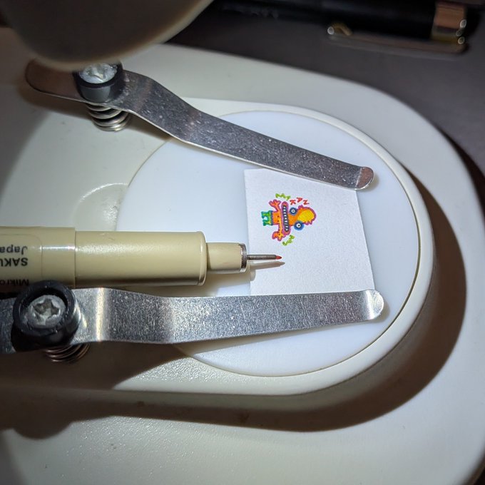 A close-up photo showing a Sakura Pigma Micron pen held by a precision tool/clamp about to color or examine a tiny colorful illustration labeled "ZATAR" on a small piece of paper.