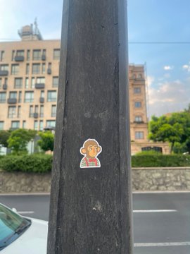 A small sticker of a cartoon character with brown curly hair is affixed to a black metal pole, with city buildings and trees visible in the background.