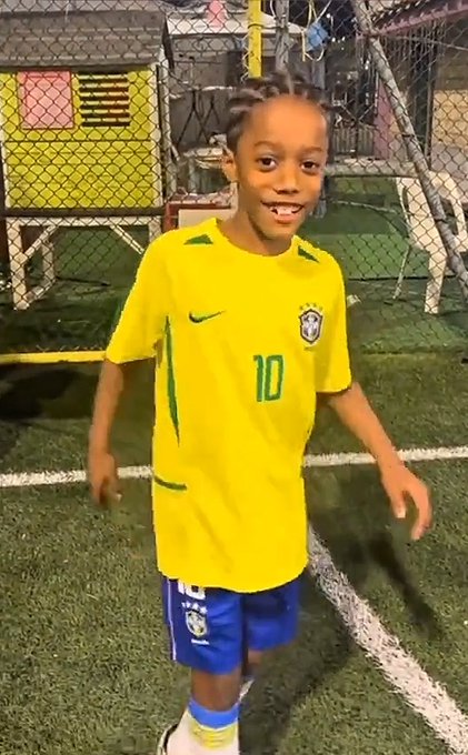 A young boy in a yellow Brazil national football jersey (#10) and blue shorts smiling on an indoor soccer field.