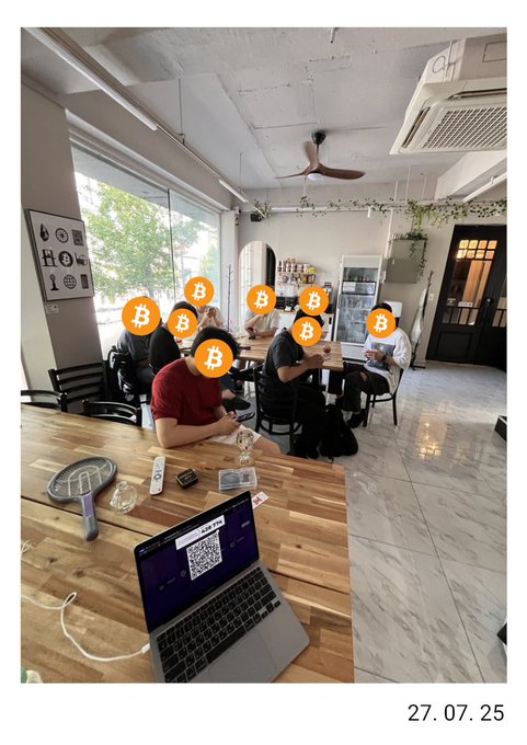 A photo dated 27.07.25 showing several people working on laptops at a wooden table in a cafe, with their faces obscured by Bitcoin logos and a QR code visible on one laptop screen.