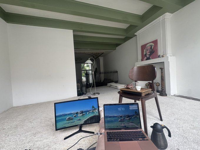 A spacious room with green ceiling beams and white walls, featuring two monitors displaying coastal scenes, a wooden chair, and a Marilyn Monroe portrait above a fireplace.