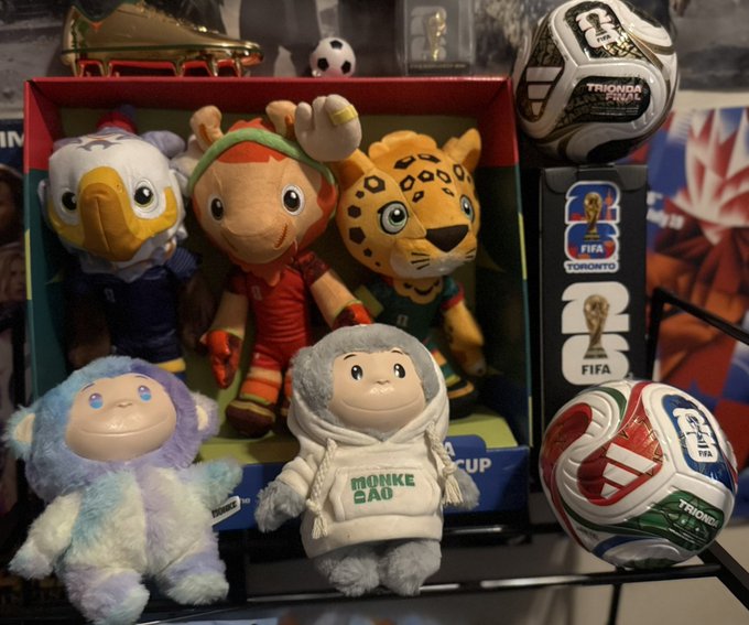 A display of World Cup memorabilia including plush mascots (an eagle, a boy, a jaguar, and yetis), soccer balls branded with "Trionda" and "FIFA World Cup 26 Toronto," and a "FIFA World Cup Trophy Toronto" figurine.