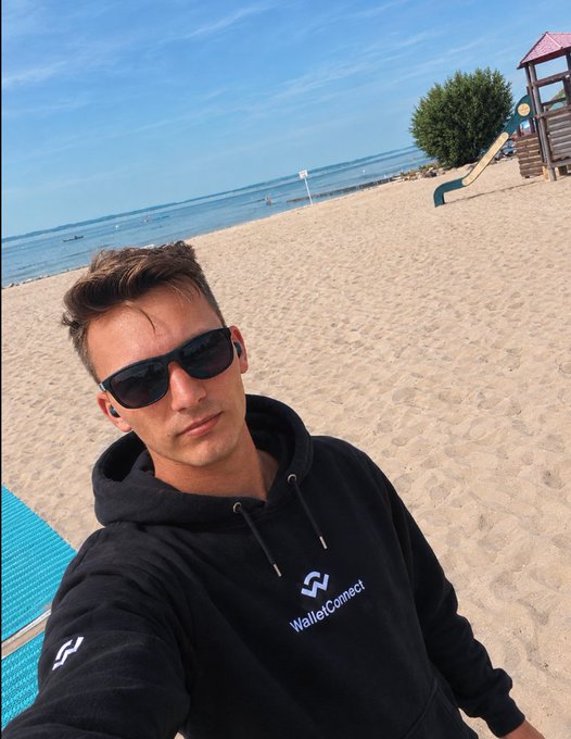 A man wearing sunglasses and a black 'WalletConnect' hoodie takes a selfie on a sandy beach with the ocean and a playground in the background.