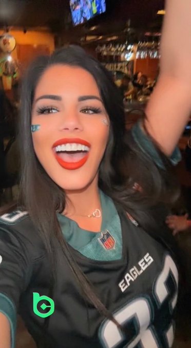 A woman with dark hair, red lipstick, and team-colored face paint is celebrating enthusiastically, wearing a Philadelphia Eagles jersey with "EAGLES" visible on the front, showing her excitement for the first game of the season in support of the Eagles and ticker $BC.