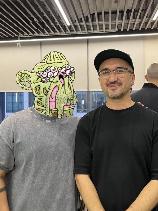 A man wearing glasses and a black cap stands next to a gray t-shirt displaying a grotesque, melting cartoon character design with multiple eyes.