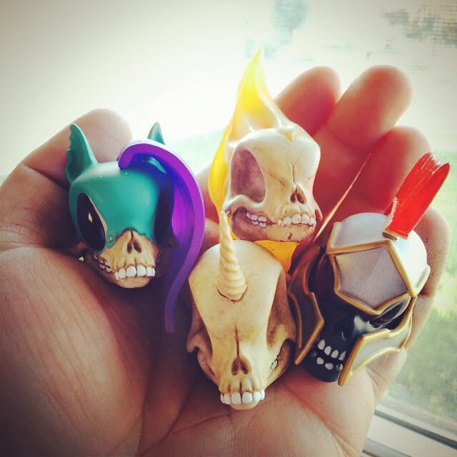 A hand holding a collection of four small, decorative skull figurines, each featuring unique designs such as horns, horns with a flame, and a ninja-like mask.