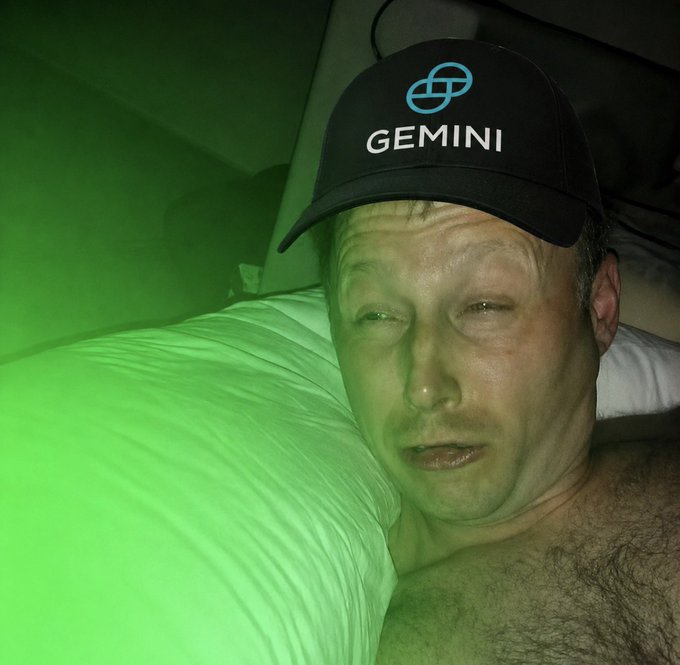 A man wearing a black "GEMINI" cap lies against a white pillow, bathed in green lighting that gives his skin an unusual tint, seemingly expressing exhaustion or illness despite his upbeat "gm" greeting to crypto friends. The Gemini logo and name reference the cryptocurrency exchange, tying into his post about bullish crypto market predictions.