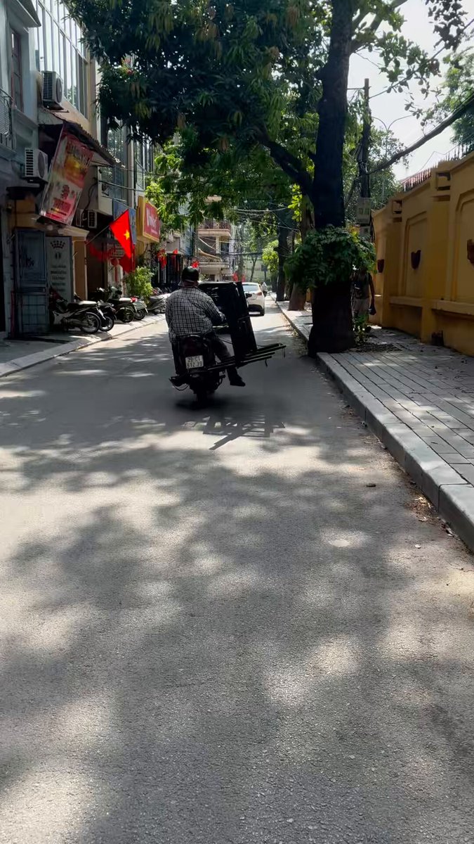 The image shows an old man riding a motorcycle down a street, with trees lining both sides of the road. The photo appears to have been taken from behind the motorcycle, giving a clear view of the rider as he moves through the scene. In the background, there are buildings and signs that add context to the location. The post accompanying the image provides additional information about the street's name or any other relevant details.