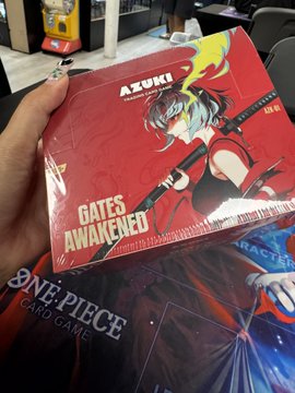 A hand holds an Azuki "Gates Awakened" booster box featuring anime-style artwork and Japanese text, with a One Piece Card Game playmat visible beneath it on a desk. The image illustrates the post about opening an Azuki box, likely a TCG booster pack product, to check for valuable pulls.