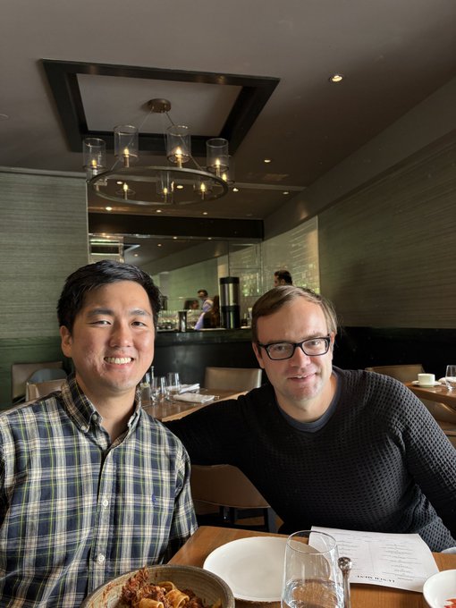 Two men pose for a photo at a restaurant, smiling at the camera, with a pasta dish, empty wine glass, and a menu (partially showing "LUNCH") on the table in front of them—appearing to capture the casual lunch meet-up referenced in the post with @vnovakovski about $LIT.