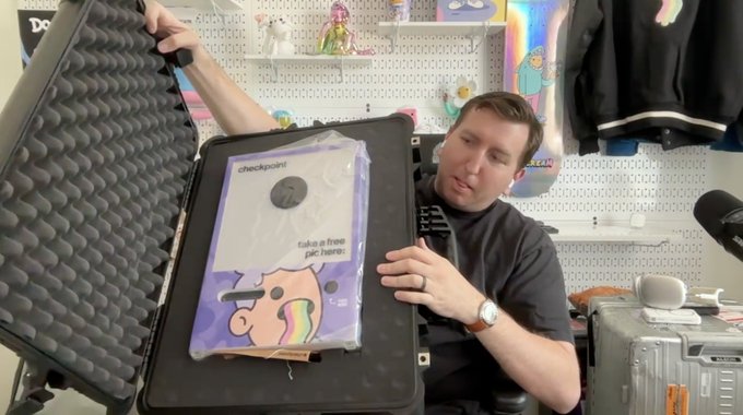 A man in a dark t-shirt sits in front of a pegboard wall, opening a black protective case to reveal a 'checkpoint' item in plastic packaging that reads 'take a free paci here' with a cartoon bear graphic.