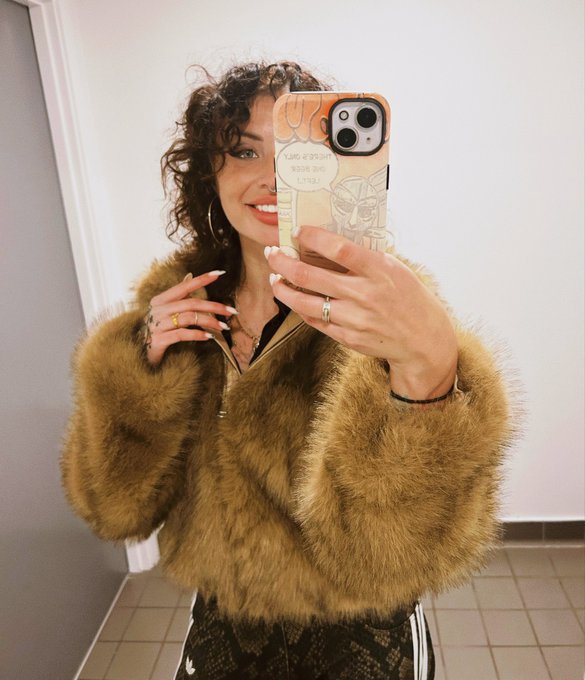 A woman with curly dark hair taking a mirror selfie, wearing a tan/brown faux fur jacket, multiple rings, and a leopard print bottom.