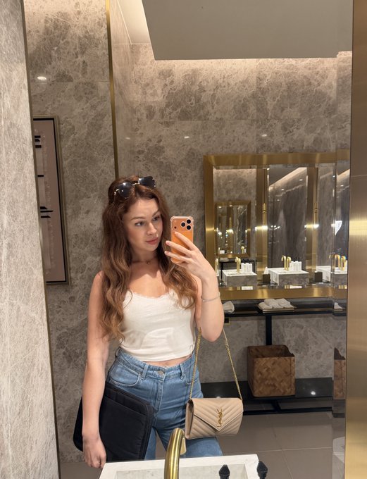 A woman takes a mirror selfie in a marble bathroom, holding an orange phone and wearing a white tank top and jeans.