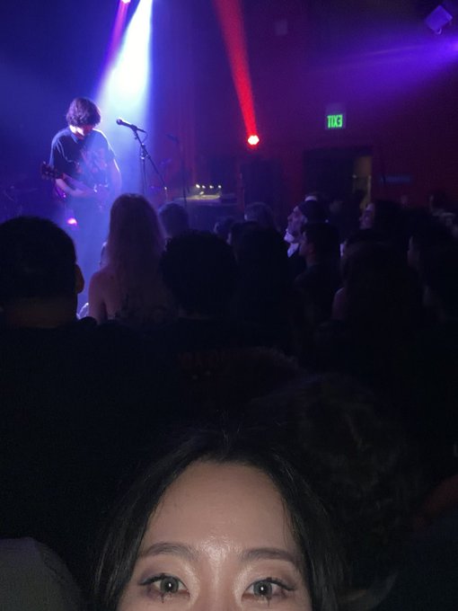 A dimly lit concert venue where a musician plays guitar on stage under blue and red lights, with a crowd of attendees visible in the foreground and a partial view of a person's face at the bottom of the frame.