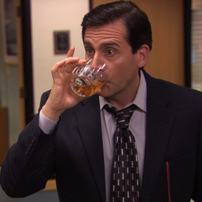 A scene from The Office showing Steve Carell as Michael Scott wearing a dark suit and patterned tie, taking a drink from a clear glass of amber liquid (whiskey).