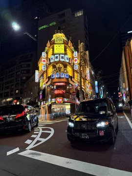 A nighttime street scene in Japan featuring a black taxi in the foreground and the brightly lit Kabukicho area with its iconic illuminated signs.