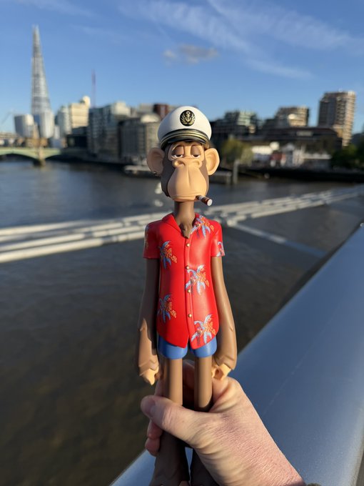 A hand holds a Bored Ape Yacht Club figurine dressed in a red Hawaiian shirt and sailor hat, with the Shard skyscraper and London skyline visible in the background.