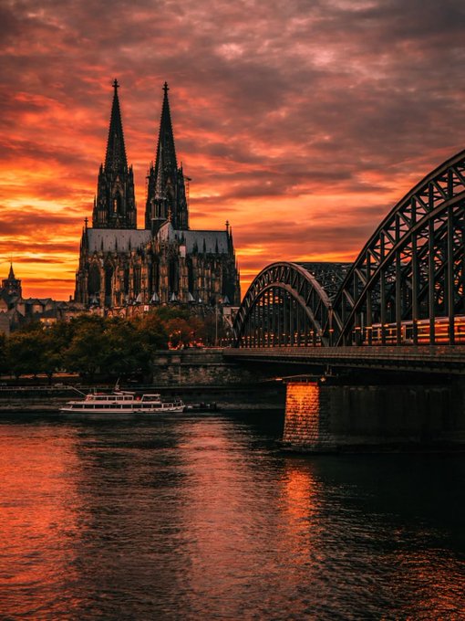 The image captures a beautiful scene at night over a river, with a cathedral featuring two spires prominently visible. The photograph also includes a bridge spanning across the water below. A boat can be seen in the middle of the river. There is another boat closer to the bottom left corner of the photo, and it appears that there are multiple boats in general throughout the scene.