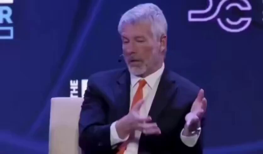 The image shows an older man wearing a suit, sitting at a podium with his hands up as if he is speaking. He has gray hair and appears to be addressing an audience. In the background, there are other people sitting behind him. 
 A text overlay in the corner of the image reads, "Bitcoin says if America buys it, its allies and enemies will have to buy it back at $10 million, $20 million, or $50 million a coin." The post suggests that buying Bitcoin can lead to significant financial gains.