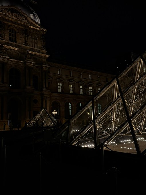 A nighttime photograph of the Louvre Museum featuring its illuminated glass pyramid and a classical building facade.
