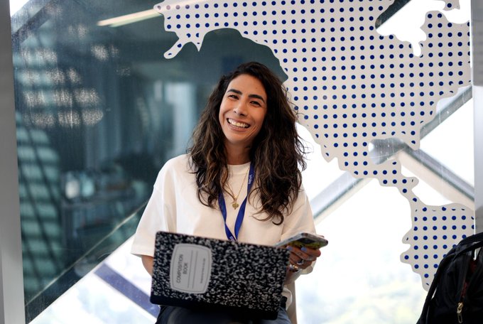 A smiling woman with long dark hair holds a composition notebook and smartphone, wearing a white t-shirt and blue lanyard, standing in front of a blue dotted world map display.