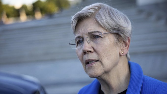 Senator Elizabeth Warren, wearing glasses and a blue blazer, speaks outdoors with a serious expression, illustrating her concerns about the Clarity Act and its potential to trigger an economic crash in the U.S.