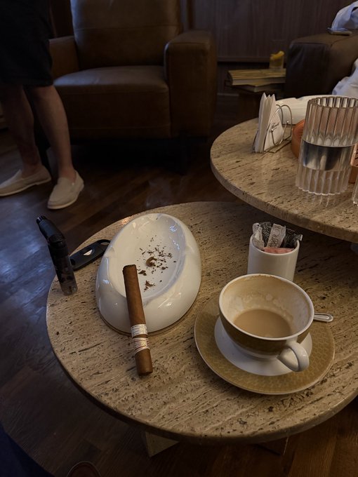 A round marble coffee table holds a white ceramic ashtray with a cigar, a cup of coffee on a saucer, a container of sugar packets, and a glass of water, set in a lounge area with a brown leather sofa.