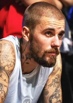 A man with a closely shaved head, full beard, and heavily tattooed arms sits in a sleeveless shirt, looking off to the side with a serious expression. The image appears alongside a post reporting that Justin Bieber is the frontrunner to perform at the 2027 Super Bowl, though the man pictured does not appear to be Bieber.