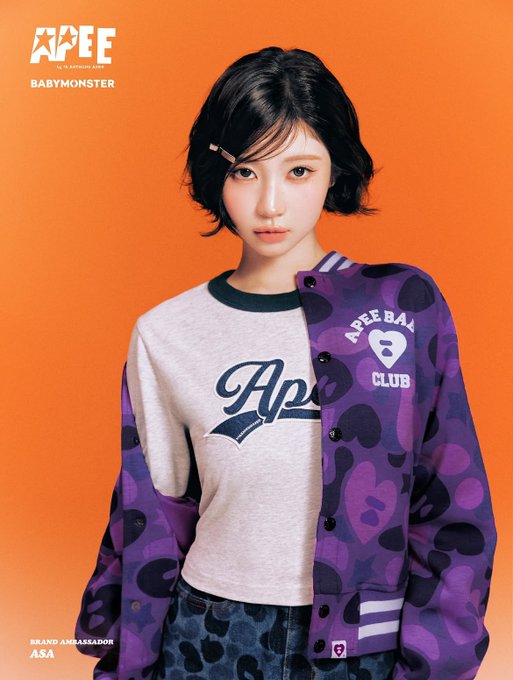 A promotional image features ASA from BABYMONSTER as the brand ambassador for the APEE Fall 26 Collection, showcasing her in a purple camouflage varsity jacket over a gray "Apee" graphic tee against a vibrant orange backdrop. The top left displays "APEE FALL 26 BABYMONSTER" while the bottom corner identifies her as "BRAND AMBASSADOR ASA."