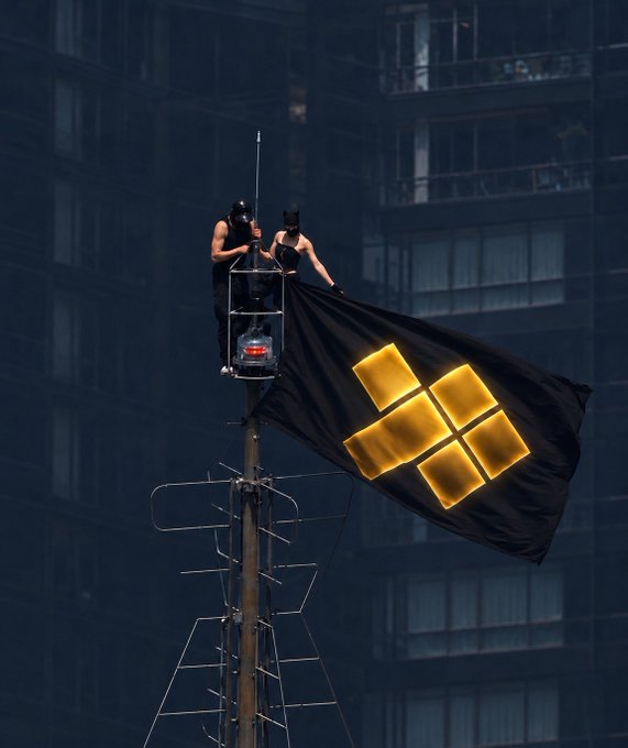 The user wants a concise caption for the image.
The image shows two people in a cherry picker or lift at the top of a pole, unfurling a large black flag with a glowing yellow logo.
The logo is four squares arranged in a cross shape. This is the logo for Binance (the "Binance Coin" or "BNB" logo, though specifically this looks like the Binance logo).
The prompt provides context: "they said gold was old... then $GGBR made it available for $4... they said crypto had no real backing, then @stakemygold paid 12% APR on it...".
The text mentions "crypto", "gold", and "$GGBR". The image shows a flag with a crypto logo (Binance). The text also mentions "stakemygold", which is a platform related to gold-backed crypto.
However, the most direct visual element is the flag being raised. The flag has the Binance logo