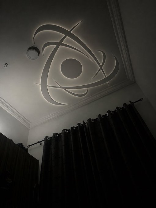 A backlit ceiling light fixture with an atom or spiral design casts dramatic shadows on the ceiling above dark curtains in a dimly lit room.