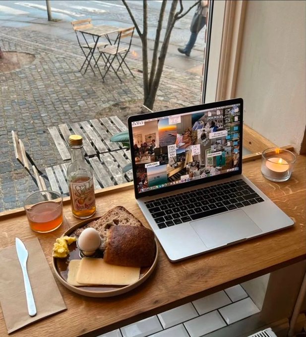 A laptop on a wooden cafe table displaying a photo collage, next to a breakfast plate with bread, egg, and cheese, a bottle, and a lit candle.