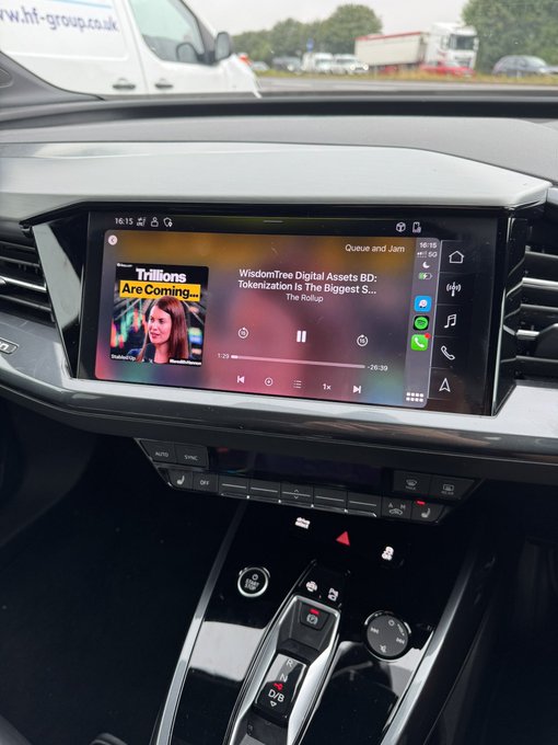 A view inside a car dashboard showing the infotainment screen playing a podcast called 'Queue and Jam' with the episode 'Trillions Are Coming...' featuring WisdomTree Digital Assets.