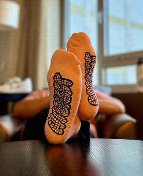 The image features a pair of feet with orange socks, placed on top of a wooden table. The socks have black lace designs on them, adding a unique touch to their appearance. The background includes a window that allows natural light into the room, creating an inviting atmosphere for the viewer.