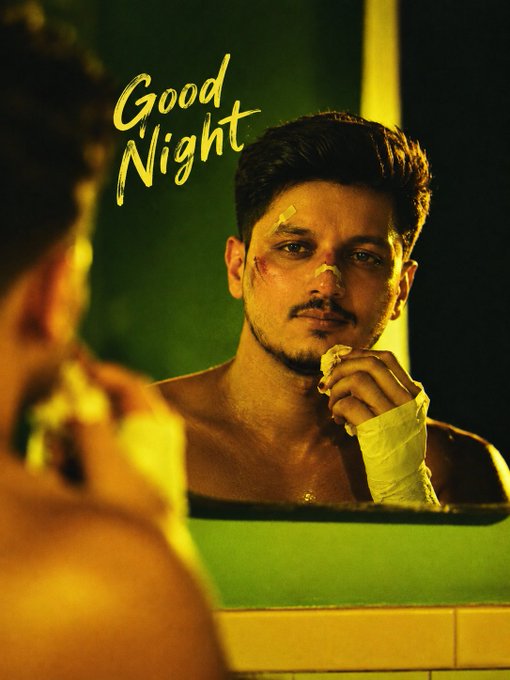 A man with facial wounds and bandages stares at himself in a bathroom mirror in a dimly lit scene, with "Good Night" written in yellow script on the wall behind him.