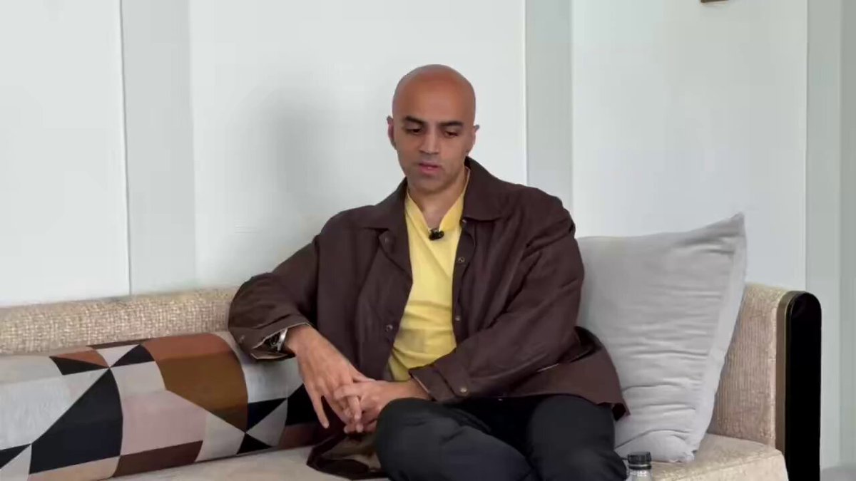 The image features a man sitting on a couch, wearing a brown jacket and a yellow shirt. He appears to be deep in thought or contemplation as he looks down at his hands. The background of the image shows a white wall with a pillow placed against it. There is also an object that resembles a bottle near the bottom right corner of the photo.