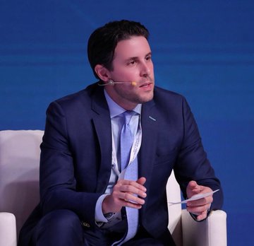 A man in a dark blue suit and light blue tie speaks into a headset microphone while gesturing with a pen, seated against a deep blue background. The image accompanies a report about Coinbase's John D'Agostino discussing 40+ countries committing to buy Bitcoin for national reserves.