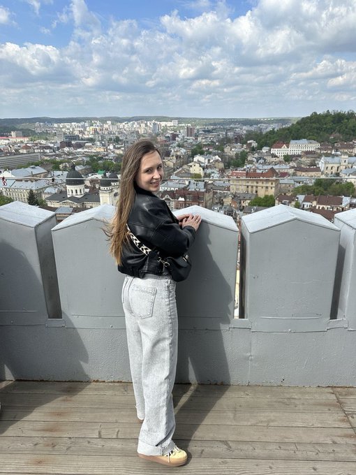 A young woman in a black leather jacket and light grey jeans stands on a wooden observation deck overlooking a city landscape.