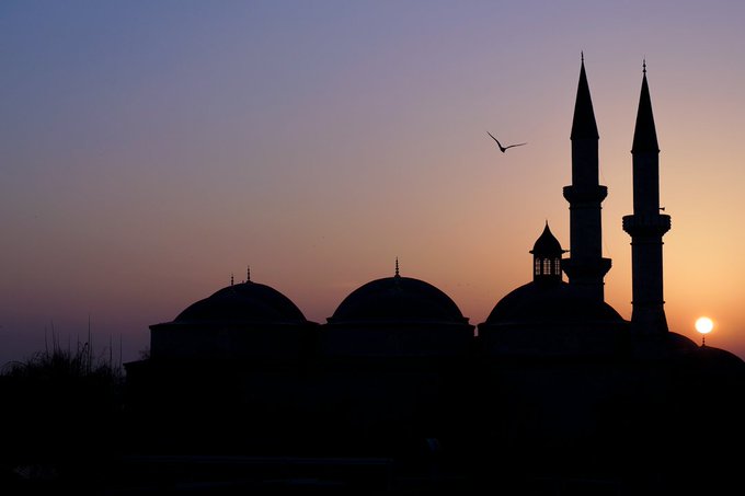 Silhouette of a mosque with domes and minarets against a twilight sky with the sun setting on the horizon and a bird in flight.