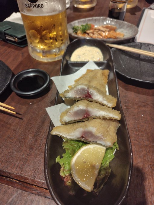The image shows a wooden table with various food items, including fish sticks or fried chicken, accompanied by dipping sauce. There are also chopsticks placed next to the plate of food. The scene appears to be set at an Asian restaurant where customers can enjoy their meal and have drinks like beer.